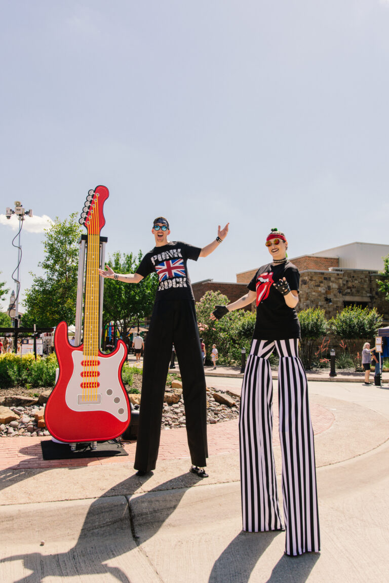 Two persons in stilts posing in front of a giant guitar
